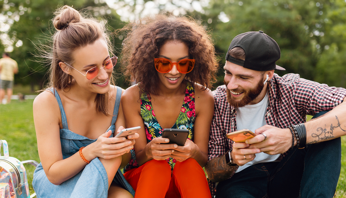 Three friends smiling and looking at their smartphones in a park.